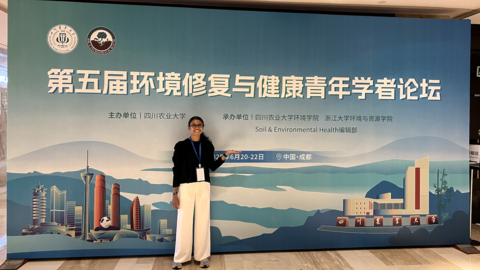 A student standing and smiling in front of a conference banner for the Soil and Environmental Health Forum in Chengdu, China.