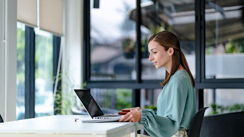 A woman works on a laptop at a bright desk near large windows.